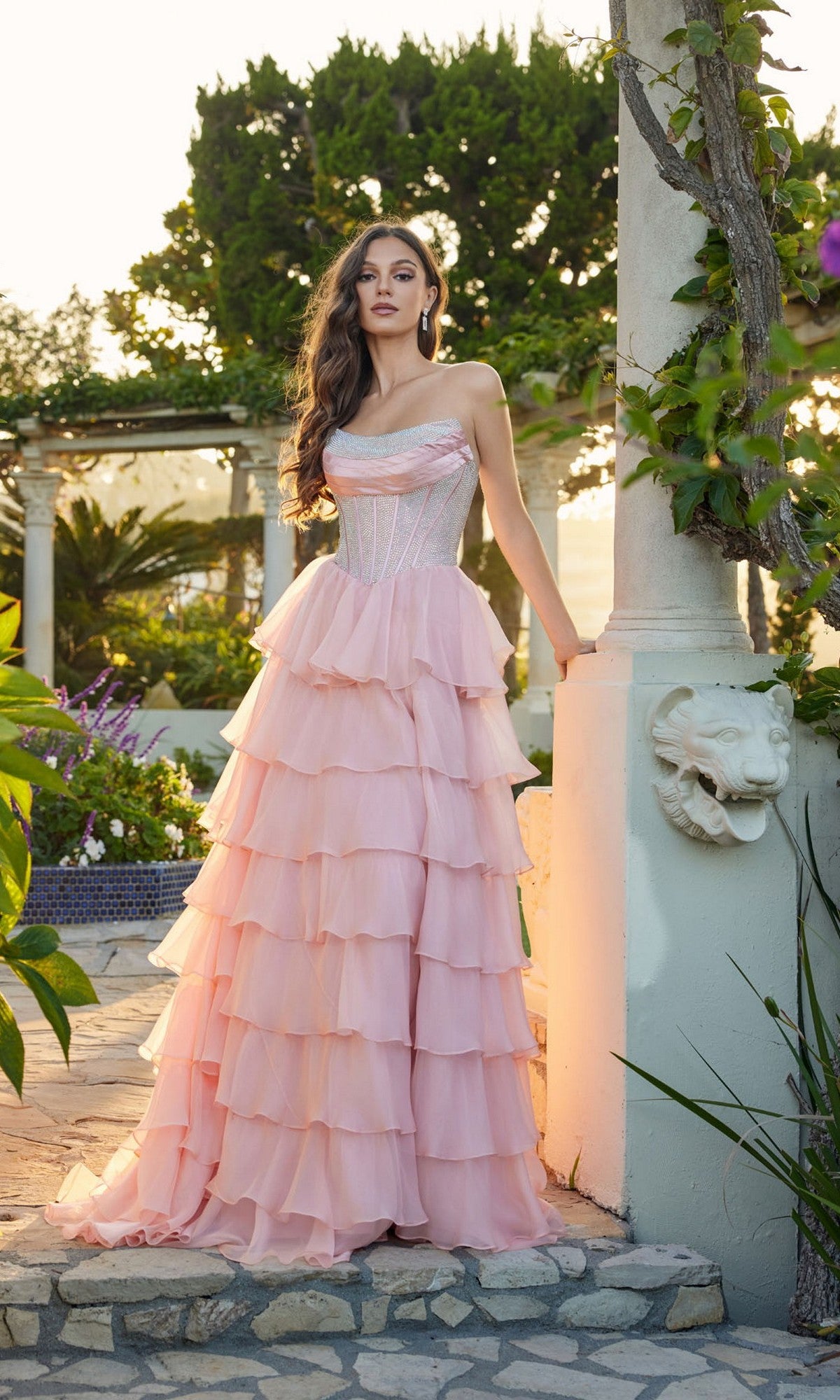 A woman in the blush Juliet JT2738S long prom dress, featuring a strapless bodice and chiffon ruffle skirt, stands outdoors by a column with greenery, sunlight streaming in.